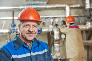 A man in blue and orange hard hat next to pipes.